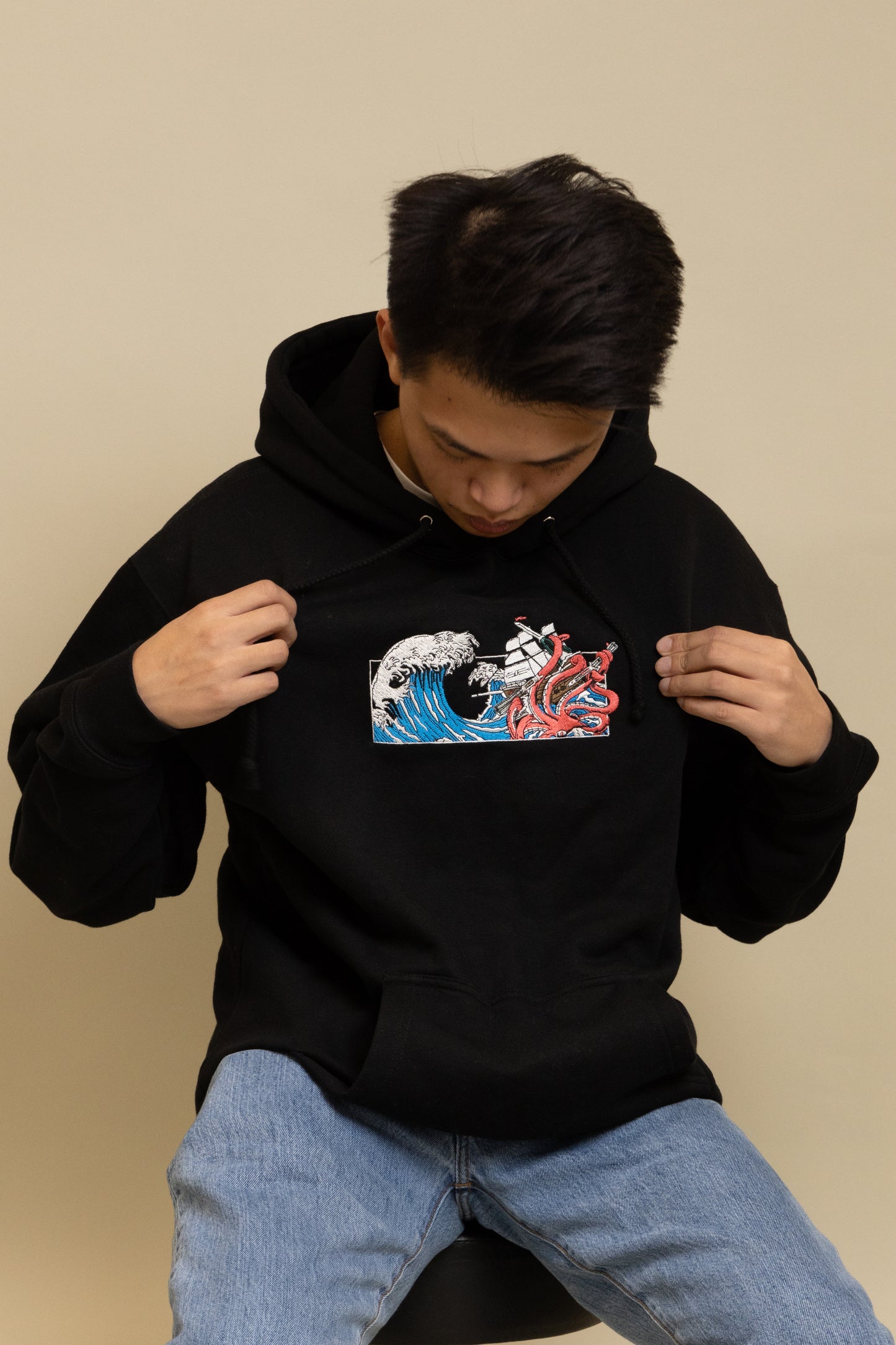 A close up view of a man wearing Black Squid Industries x Simple Stock Hoodie posing while sitting on a chair