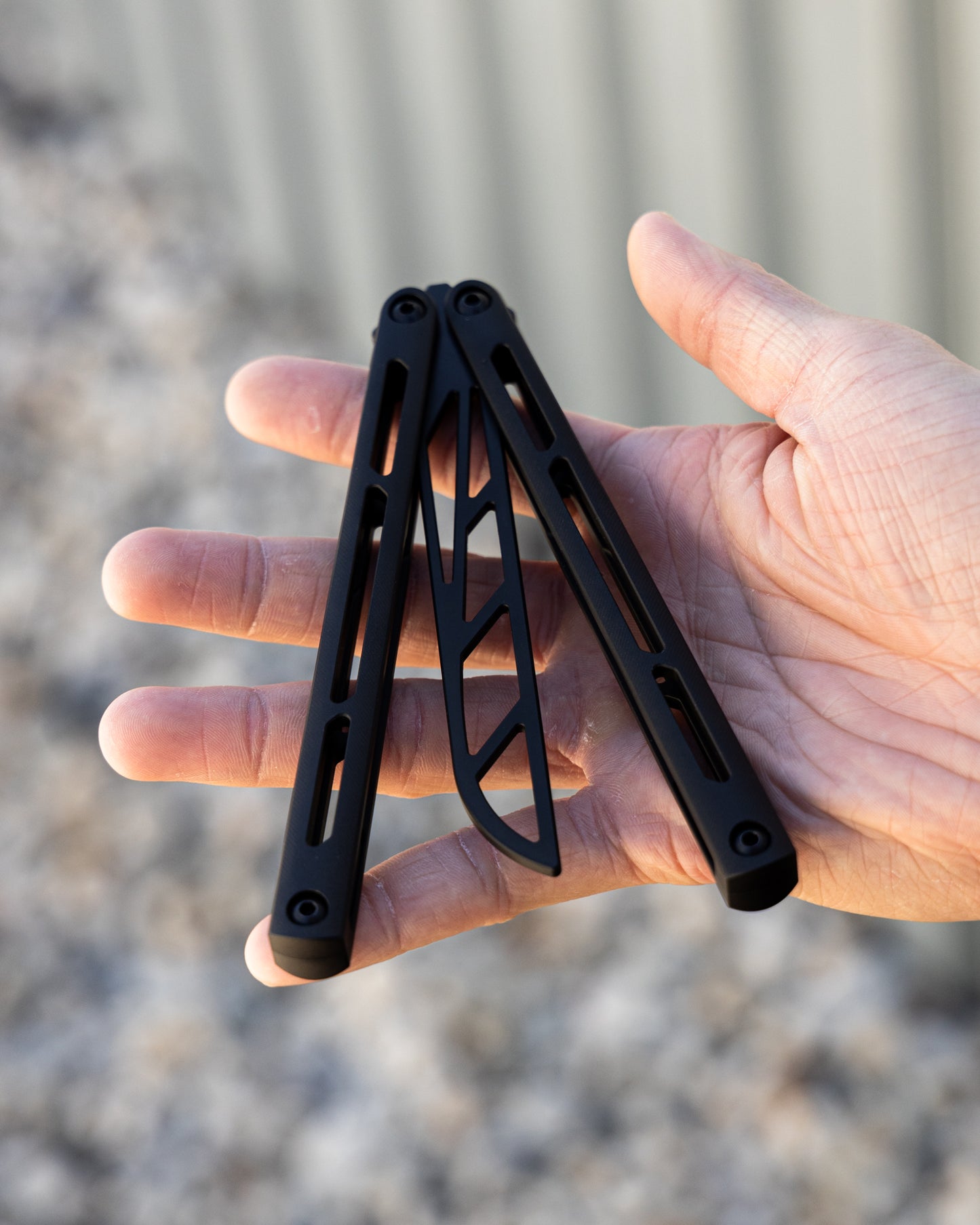 Black metal folding knife trainer held in a hand with a blurred natural background