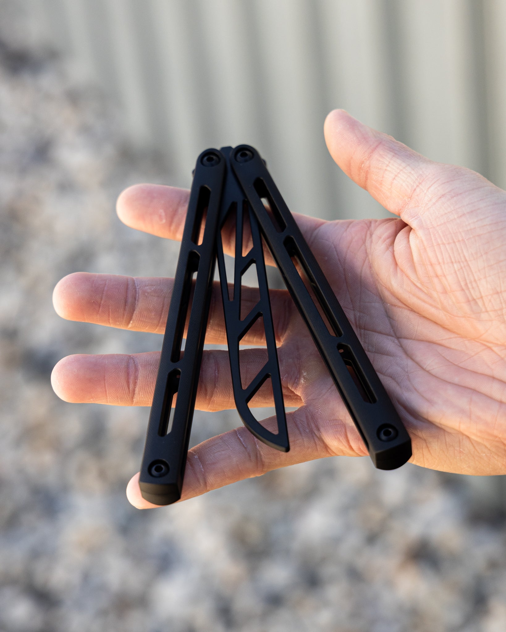 Black metal folding knife trainer held in a hand with a blurred natural background