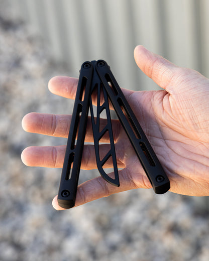 Black metal folding knife trainer held in a hand with a blurred natural background