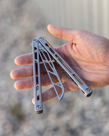Hand holding a silver balisong trainer against a blurred natural background