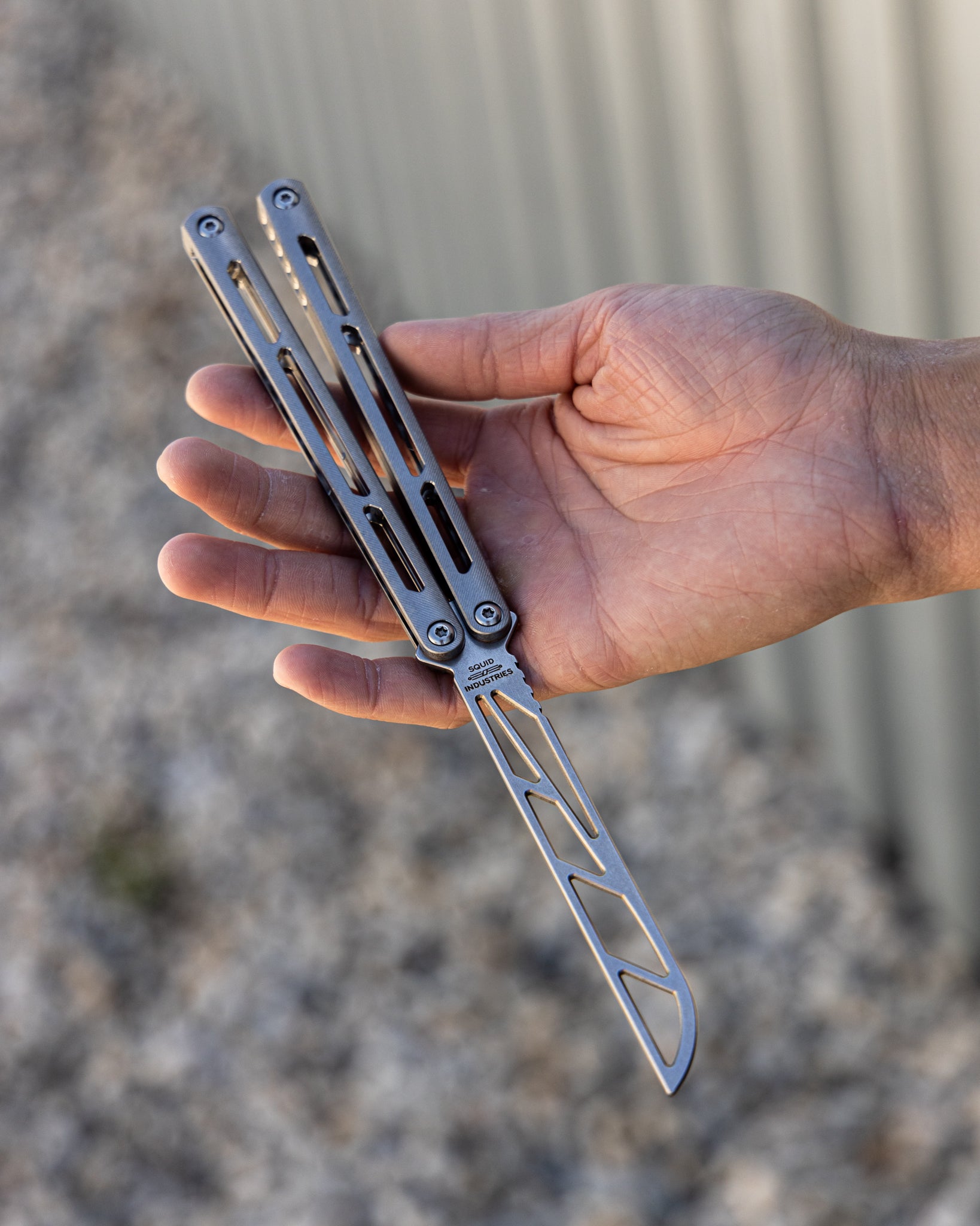 Hand holding a silver metal balisong trainer with a blurred background