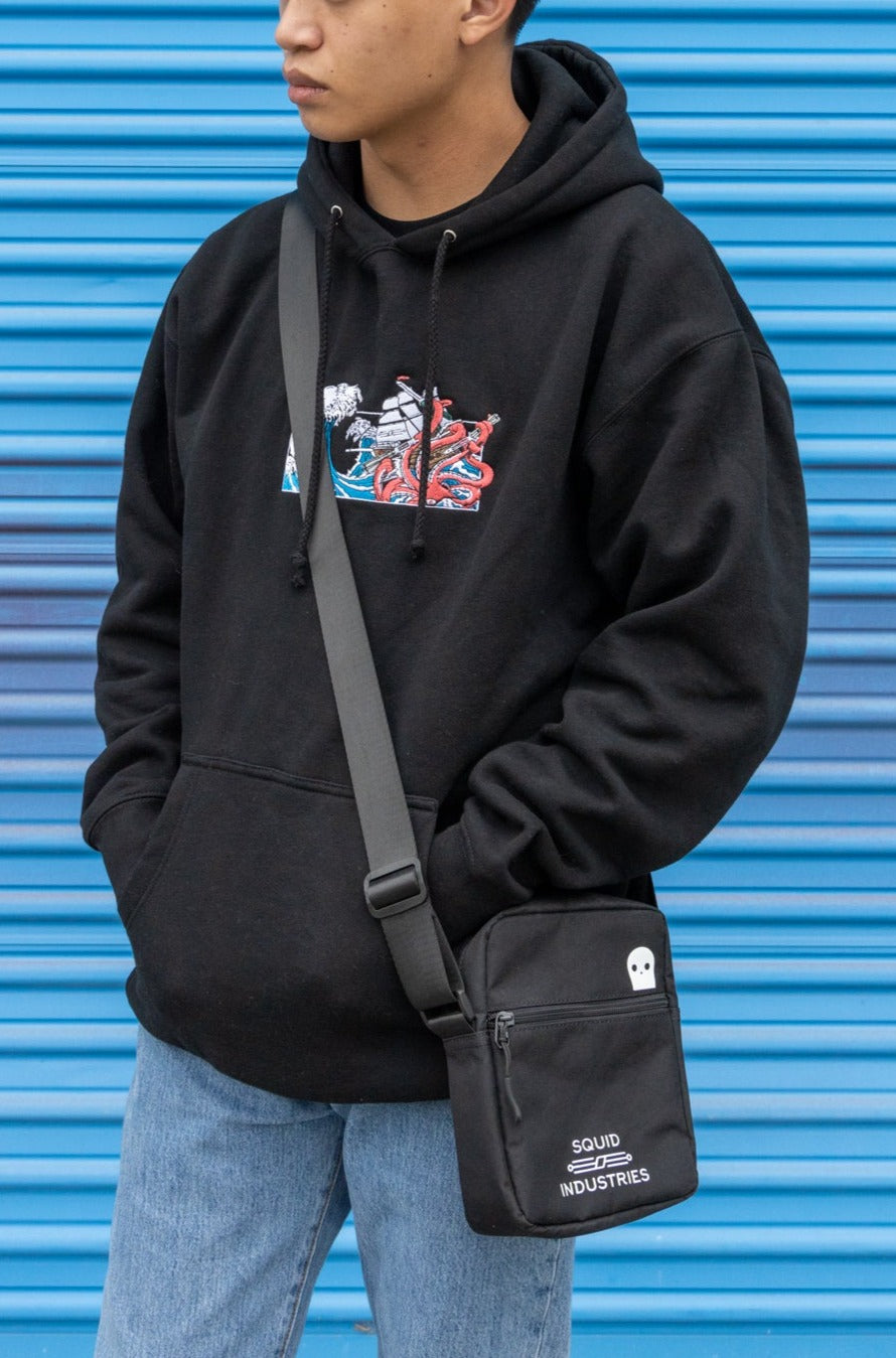 Man wearing Squid Industries x Simple Stock Black Hoodie while wearing a black Squid Industries crossbody bag against a blue background 