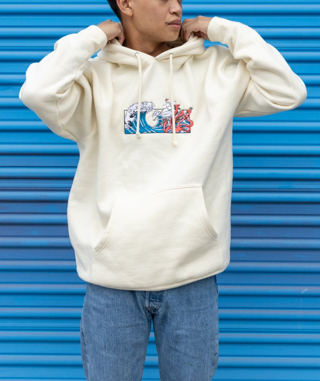 Man wearing a cream Squid Industries x Simple Stock Hoodie posing against a blue background outside